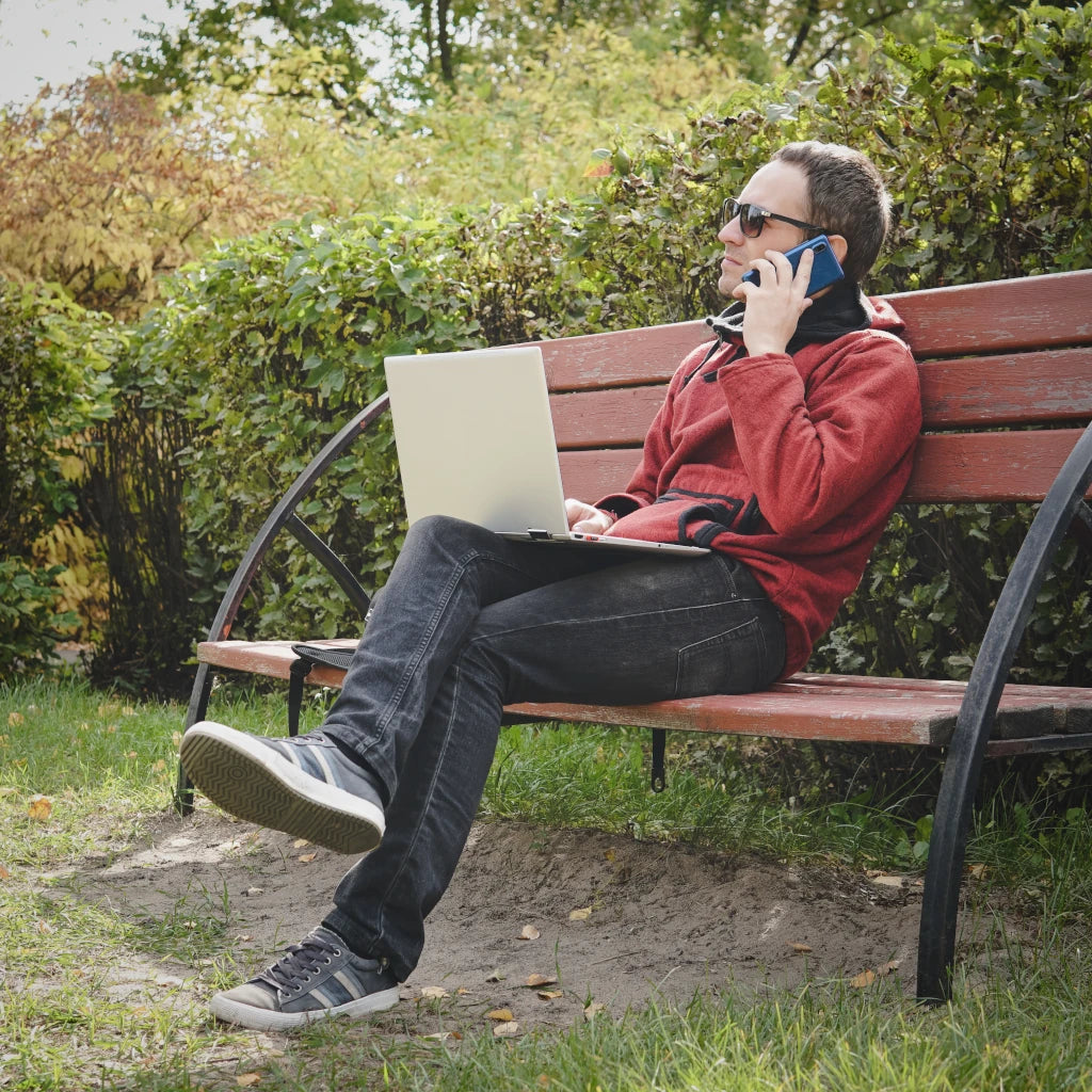 Person sitting on a park bench with a laptop and phone, surrounded by greenery.