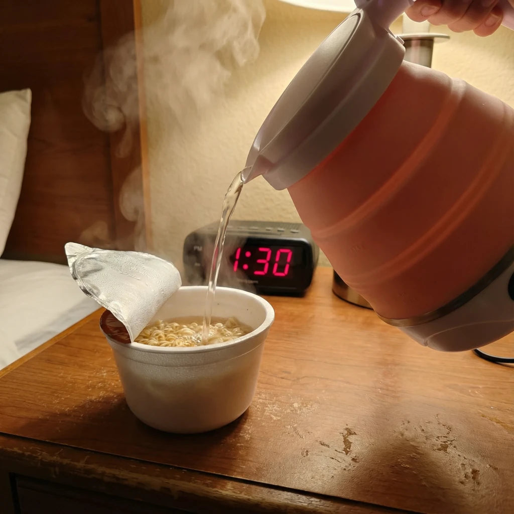 Person pouring liquid from a pink collapsible electric kettle into a white ramen bowl on a wooden hotel desk.