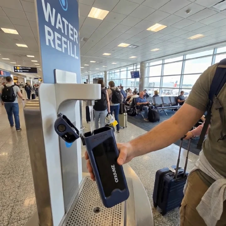 Person using a rectangular sports bottle at a refill station at an airport with 'WATER REFILL' sign.