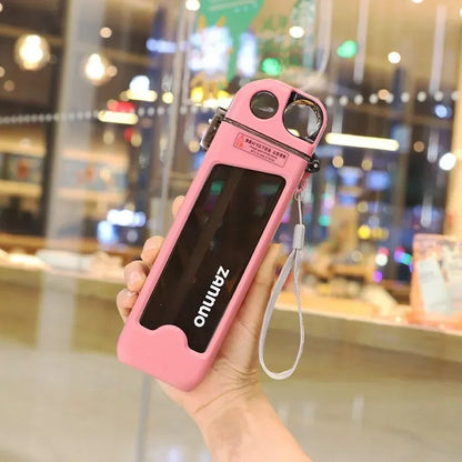 Pink square water bottle with camera lens attachment held by a hand in front of a blurred indoor background