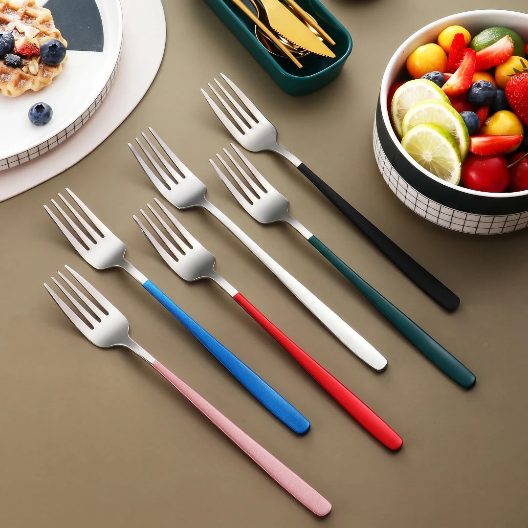 Set of forks with varying color handles  on a brown surface with a bowl of fruit and waffles in the background.