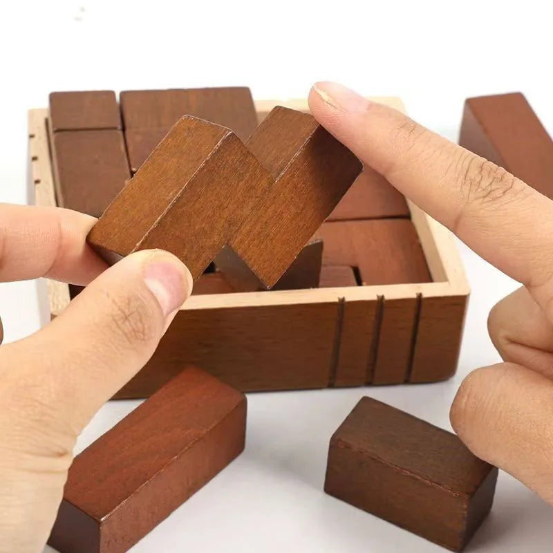 A close-up of hands holding a wooden puzzle block with the wooden box in the background and several rectangular wood pieces visible on a white background.